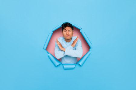 Frowning young man stands in middle of cut out hole in blue background with his hands folded crosswise showing stop gesture, advance notice concept, copy spaceの写真素材