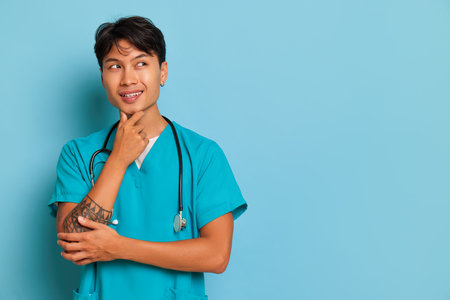 Young male nurse stands on blue background, thinking and holding his chin with one hand and smiling, professional people concept, copy spaceの写真素材