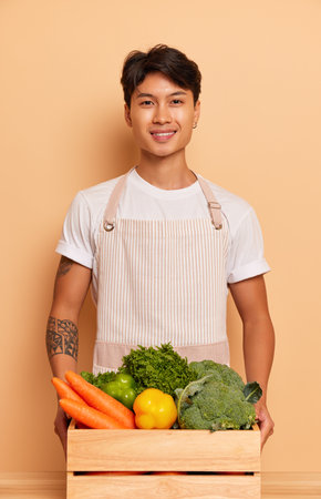 Cheerful greengrocer in white T-shirt and in apron posing inside holding box full of fresh vegetables in hands, organic food concept, copy spaceの写真素材