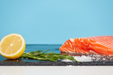 Slice of salmon with spices and lemon isolated on blue background, still-life with tasty fish fillet on black cutting board, seafood dish concept, copy spaceの写真素材