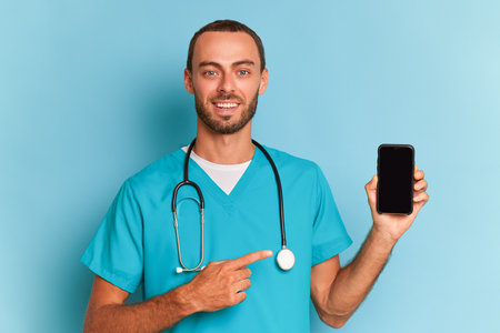 Young male medical worker holds phone screen in hand and points at it, looks at camera and smiles, wears special medical shirt, professional people concept, copy spaceの写真素材