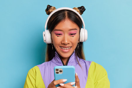 A beautiful female student in headphones stands on blue background and looks at the phone, smiles, received good message from friend, copy space, surprising news concept.の写真素材