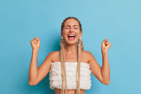 Portrait of young woman with long blond hair dreadlocks on blue background in the studio, girl stands with her hands up in joy, opened her mouth and closed eyes, happy time concept, copy spaceの写真素材
