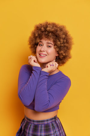 Beautiful girl in curly wig dressed in purple top and plaid skirt posing on yellow background, pressing her hands to her chest, funny moments concept, copy spaceの写真素材