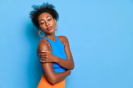 Close up of pretty black girl in glasses, stands in beautiful pose against blue wall, hugging herself closing her eyes, wears blue top, relax concept, copy spaceの写真素材