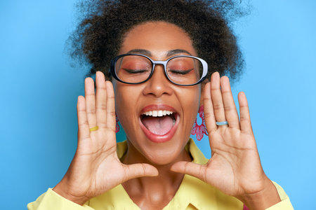 Close up shot of black girl in glasses on blue background, closing her eyes with palms at mouth and...