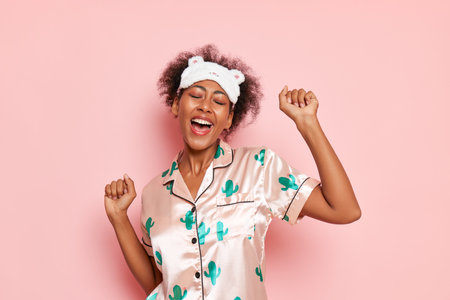 Young black pleased woman dancing against pink wall, wearing silk pyjamas and sleep mask on head, good morning concept, copy spaceの写真素材