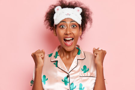 Young black woman posing against pink wall with happy expression on her face, wearing silk pyjamas and sleep mask on head, holds fists up, good morning concept, copy spaceの写真素材