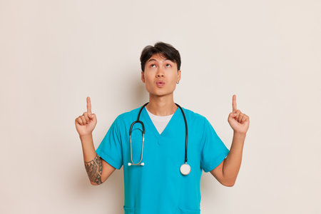 Young asian male doctor in medical uniform poses on white background, looks up and points with index fingers up, professional people concept, copy spaceの写真素材