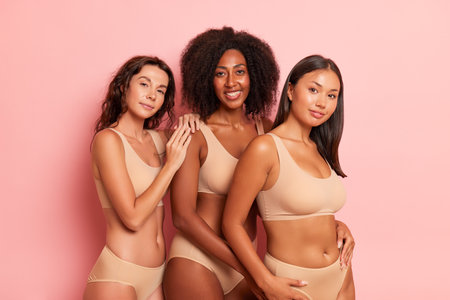 Cheerful young women in beige undergarment pose body half-turned against pink background one puts hand on shoulder of friend next to, comfortable underclothing concept, copy spaceの写真素材