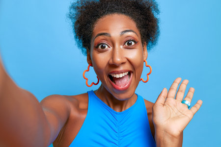 Close-up shot of pleased black girl in blue top smiling broadly while taking selfie against blue wall, happy time concept, copy spaceの写真素材