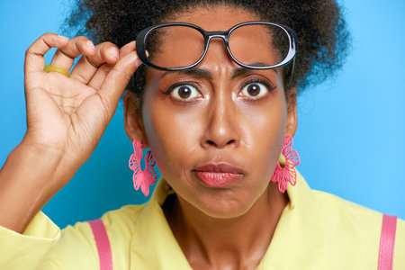 Portrait of young black woman wearing yellow shirt, close-up face on blue wall, holding glasses up on her forehead, serious moment concept, copy spaceの写真素材