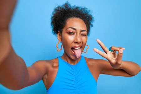 Portrait of black girl in blue top having fun while taking selfie against blue wall, puts out her tongue and shows victory sign, happy time concept, copy spaceの写真素材