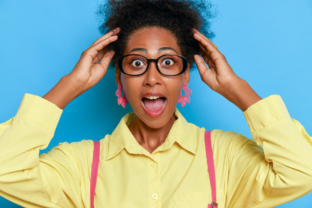 Portrait of girl with black skin and lush hair, posing in yellow shirt on blue background, holding her hands on her head, excitement concept, copy spaceの写真素材