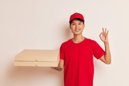 Cheerful chinese guy holding box with pizza in one hand, shows ok sign with other hand, wearing red cap and red t-shirt, fast delivery concept, copy spaceの写真素材