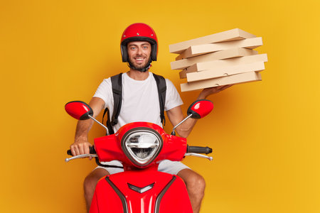 Bearded young man dressed in white t-shirt and red helmet, riding red scooter, holds pack of pizza boxes in one hand, fast drive concept, copy spaceの写真素材