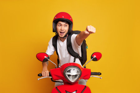 Chinese guy in red helmet rides red scooter posing on yellow background, one hand clenched into fist and extended forward, food delivery concept, copy spaceの写真素材