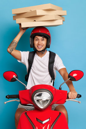 Chinese boy sits on his red scooter on blue background, holding stack of pizza boxes on his head, food delivery concept, copy spaceの写真素材