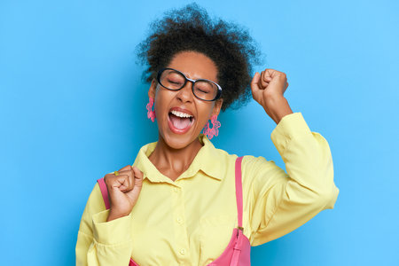 Portrait of happy girl with black skin posing on blue background, dressed in yellow shirt, holding her hands up, excitement concept, copy spaceの写真素材