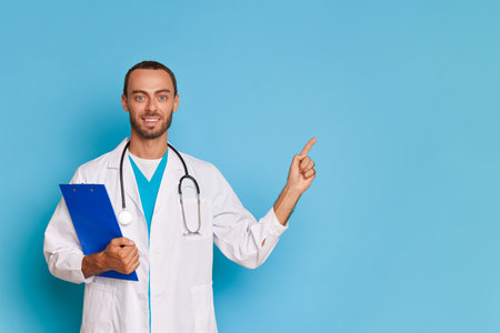 Portrait of a smiling male doctor with stethoscope holding clipboard and pointing finger up isolated over blue backgroundの写真素材
