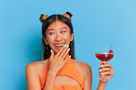 Portrait of a beautiful young asian woman in orange swimsuit holding cocktail and looking at camera isolated over blue backgroundの写真素材