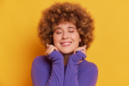 Close up portrait of young woman with Afro hairstyle wearing purple sweater, posing isolated over yellow background.の写真素材