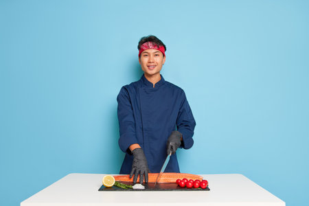 Oriental guy in blue chefs uniform and black gloves stands at cooking table in studio and slices salmon fillet, tasty food concept, copy spaceの写真素材