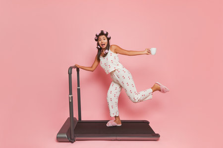 Girl in curlers on long black hair dressed in silk pajamas doing morning gymnastics, on treadmill with cup of coffee in her hand, healthy lifestyle concept, copy spaceの写真素材