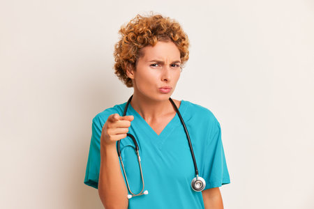 Curly female doctor standing at white backdrop, wearing blue medical uniform with stethoscope on her neck, health worker concept, copy spaceの写真素材