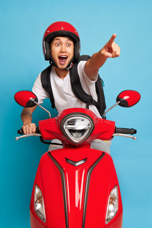 Happy guy in red helmet posing on red scooter, one hand on handle bar, another points ahead, fast drive concept, copy spaceの写真素材