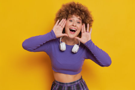 Portrait of pretty girl with afro hairstyle dressed in purple top with long sleeves posing on yellow background, satisfied smile, hands raised to face like megaphone, happy time concept, copy spaceの写真素材