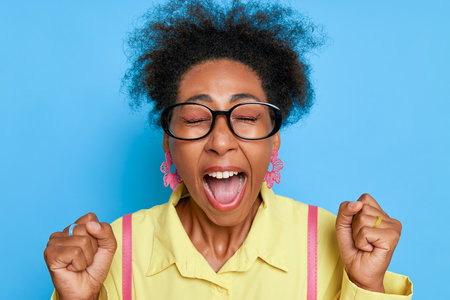 Portrait of black woman with expression of joy on her face on blue background, closed eyes, open mouth, fists up, happy moment concept, copy spaceの写真素材