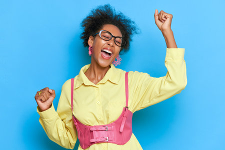 Young black woman in yellow shirt on blue background dancing with closed eyes holding arms up, expression of joy on her face, closed eyes, open mouth, happy life concept, copy spaceの写真素材