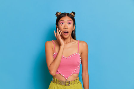 Japanese woman in pink short top poses against blue background, holds one hand near her face, opens mouth in surprise, amazing case concept, copy spaceの写真素材
