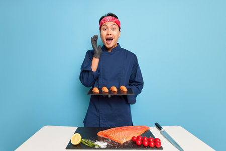 Japanese guy in blue cook uniform and black gloves stands at cooking table in studio and holds plate with sushi, tasty food concept, copy spaceの写真素材