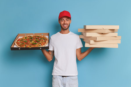 Delivery guy in red cap and white t-shirt stands with pile of pizza boxes in one hand and large pizza in another hand, fast delivery concept, copy spaceの写真素材