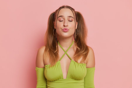 Young woman close-up dressed in green longsleeve top posing against pink background, closes her eyes and sends air kiss, perfect mood concept, copy spaceの写真素材