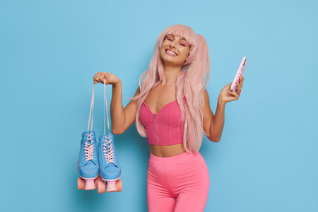 Pretty woman posing on blue background in pink clothes with pink hair, holding roller-skates and phone in hands and smiles, fashion concept, copy spaceの写真素材