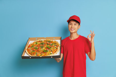 Cheerful boy with box with large pizza in one hand on blue background shows ok sign with other hand, wearing red cap and red t-shirt, fast delivery concept, copy spaceの写真素材