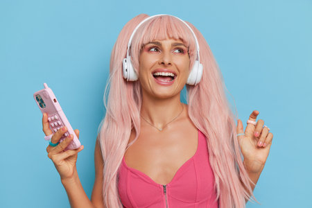 Attractive woman with pink hair and headphones wearing style pink top posing on blue background with phone in hand and smiles, vintage fashion concept, copy spaceの写真素材