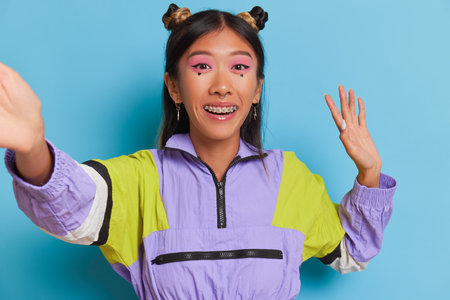 Thai girl with dental brackets and funny makeup in violet-colored sweatshirt on blue background taking selfie smiling and waving hand, good mood concept, copy spaceの写真素材