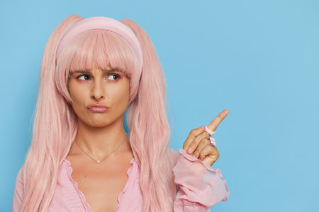 Closeup of funny girl with pink hair wearing Barbie style pink shirt posing on blue background with index finger up, vintage fashion concept, copy spaceの写真素材