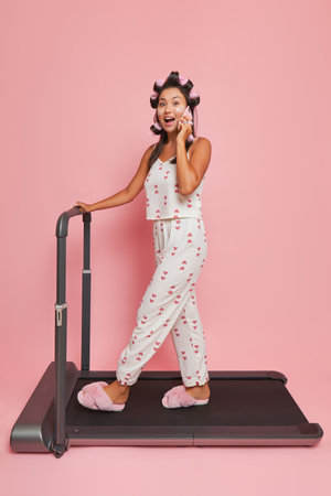 Young woman posing on pink background in white silk pajamas, doing morning exercises on treadmill while talking on phone, lifestyle concept, copy spaceの写真素材