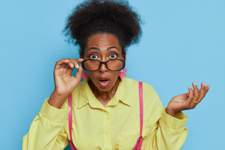 Close up coloured girl dressed in yellow shirt poses on blue backdrop, surprised look, touches her glasses with one hand, mouth opened, copy spaceの写真素材