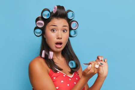 Young brunette woman in red top posing on blue backdrop, doing nails, curling hair, rushing to work, beauty care concept, copy space.の写真素材