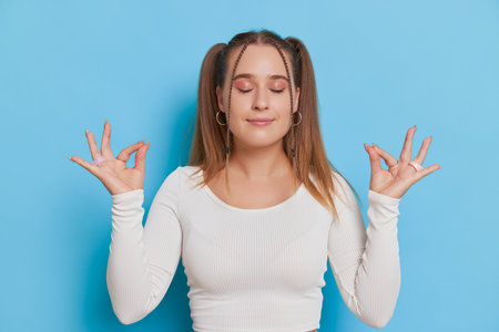 Pretty girl in white top with two ponytails stands on blue background and meditates, her eyes closed, her hands folded yoga mudra gesture, relax time concept, copy spaceの写真素材