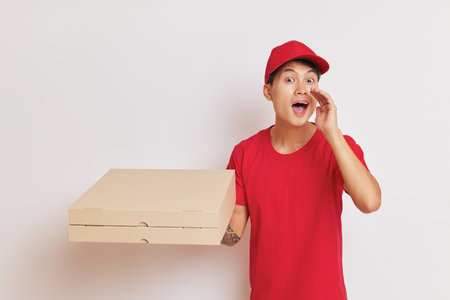Delivery guy dressed in red cap and t-shirt on white background holds pizza boxes in one hand and puts other hand to his mouth, fast delivery concept, copy spaceの写真素材