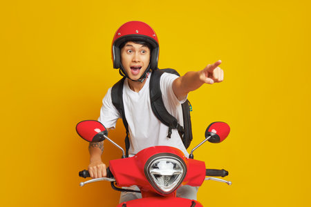 Oriental guy in red helmet sits on red scooter in yellow studio, one hand on handle bar another hand points ahead, fast drive concept, copy spaceの写真素材