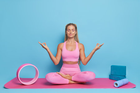 Slender girl in pink top and leggings sits on pink yoga mat against blue background, legs bent, symbolic gesture of fingers mudra, lifestyle concept, copy spaceの写真素材