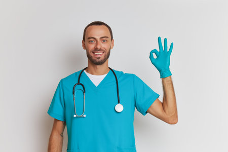 Portrait of happy male doctor on white background in medical uniform, smiles and shows ok sign with one hand, professional people concept, copy spaceの写真素材